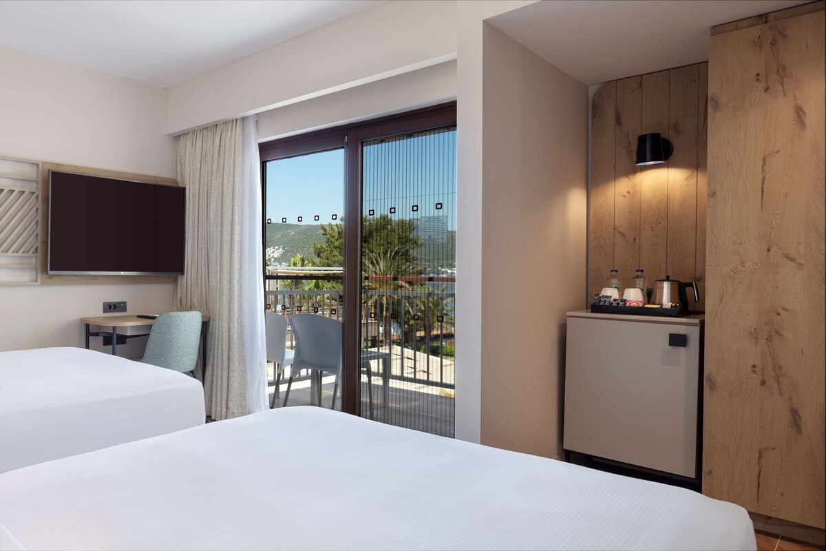Habitación de hotel con dos camas, televisión, una mesita con sillas, balcón con vistas y minibar.
