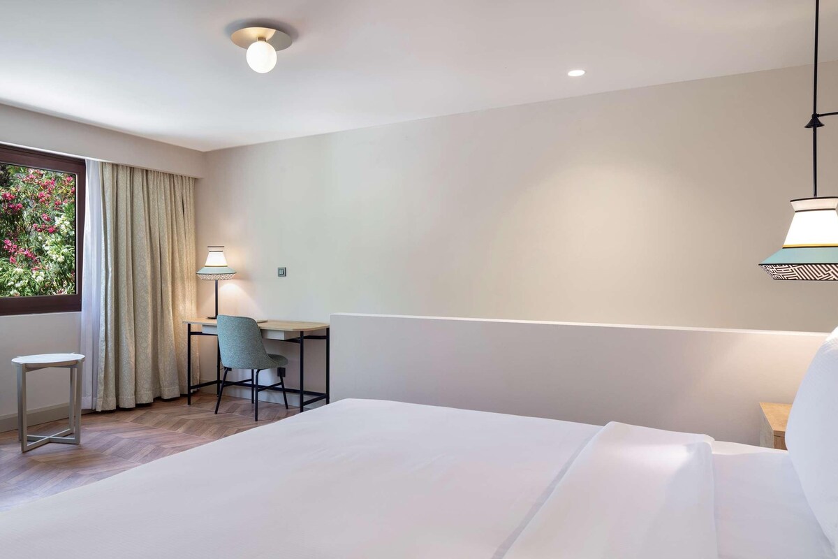 Habitación de hotel con una cama grande, un escritorio, una silla y vistas a zonas verdes por la ventana.