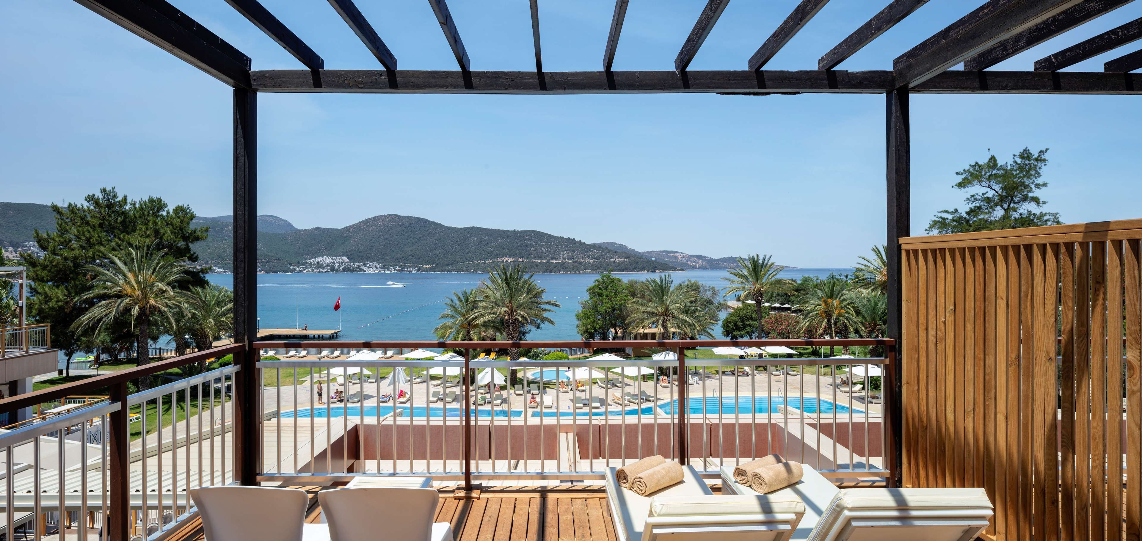 A balcony with a view of a pool and the sea, wooden furniture, and a pergola structure.