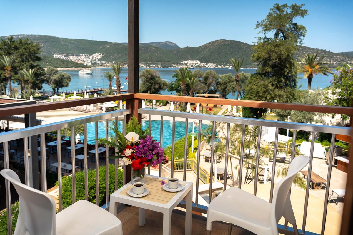 A balcony with a table and chairs, overlooking a pool and a scenic view of the sea and hills.