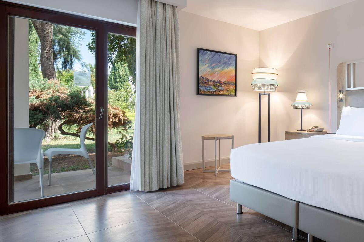 A hotel room with a large bed, a desk, a chair, and a view of the outdoors.