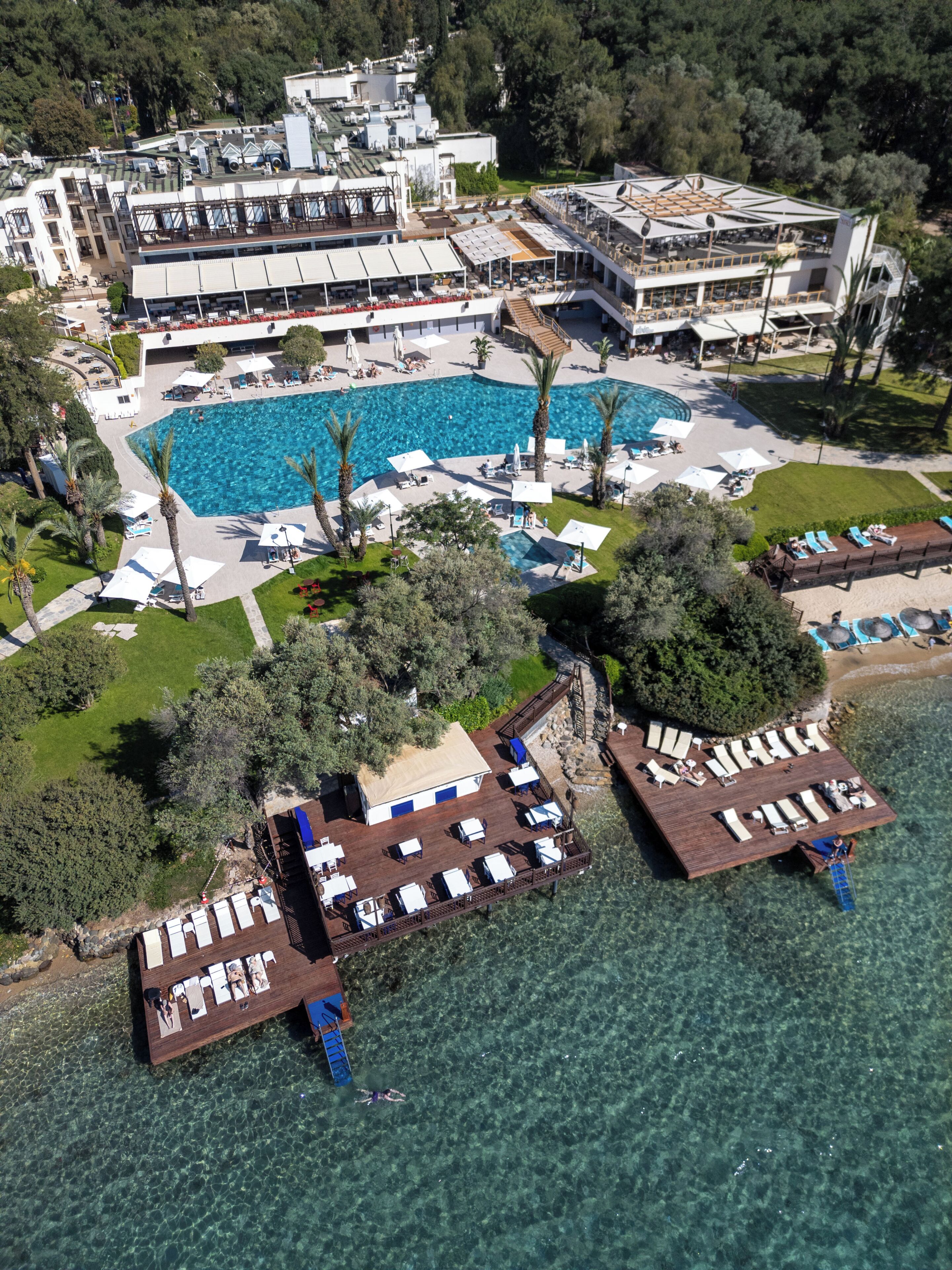 Vista aérea de un complejo turístico con una gran piscina, varias zonas de descanso y un muelle que se adentra en el agua.