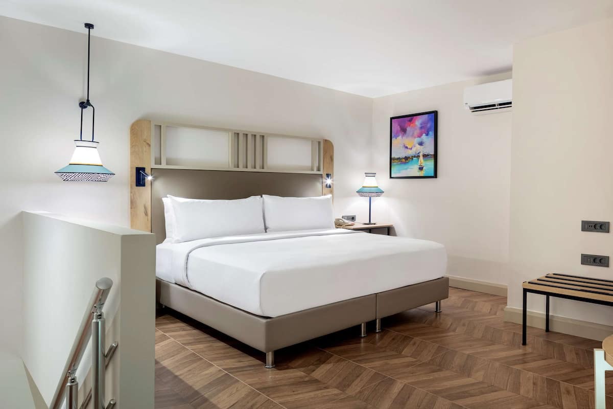 A modern hotel room with a bed, bedside lamps, a wooden wardrobe, a small table, and a chair.