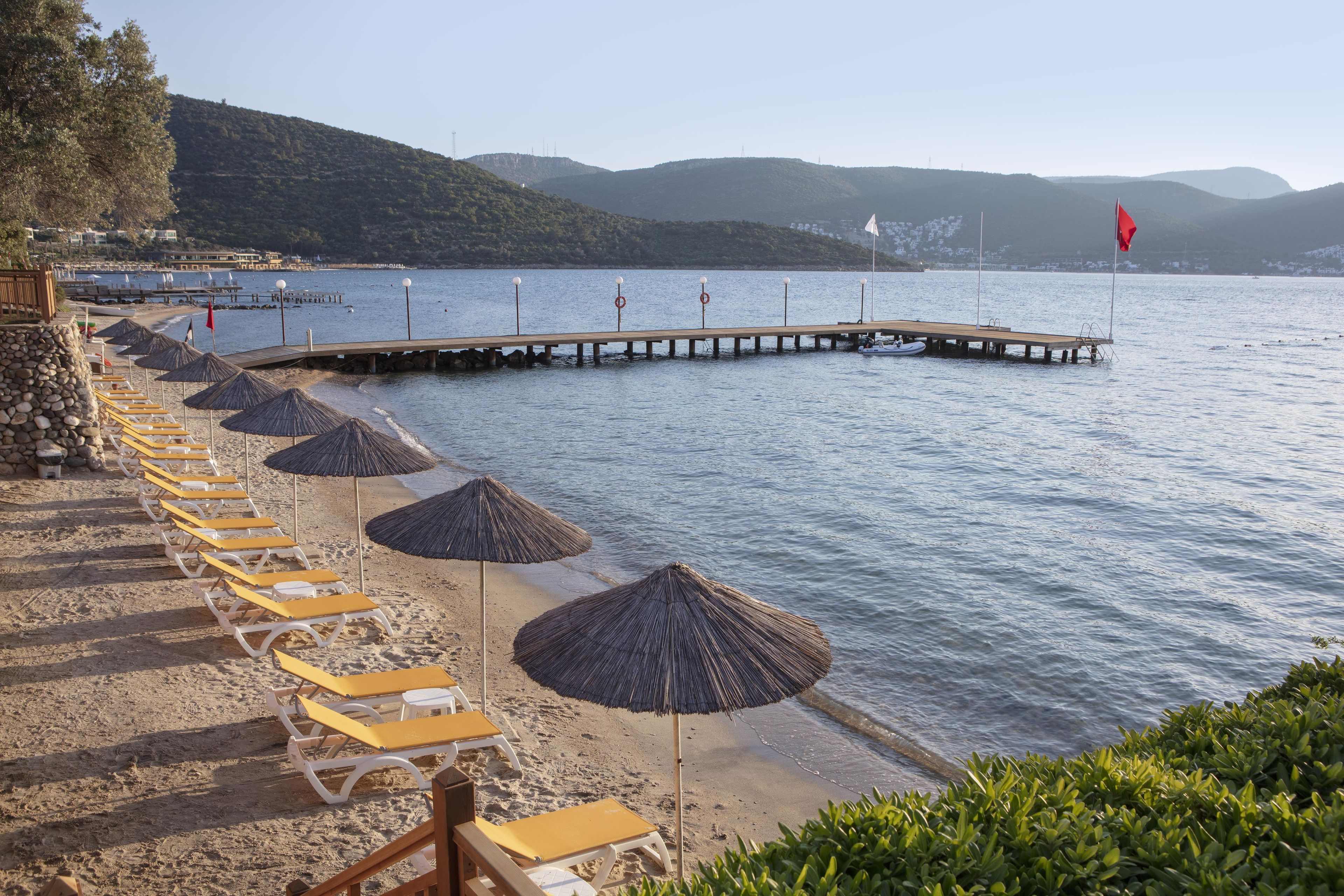 Ein Strand mit Reihen von Liegestühlen und Reetdachschirmen, ein hölzerner Steg, der sich über ruhiges Wasser erstreckt, und eine gebirgige Kulisse.