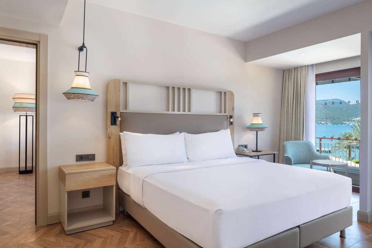 Habitación de hotel con cama, mesitas de noche, escritorio, silla y balcón con vistas al mar y colinas.