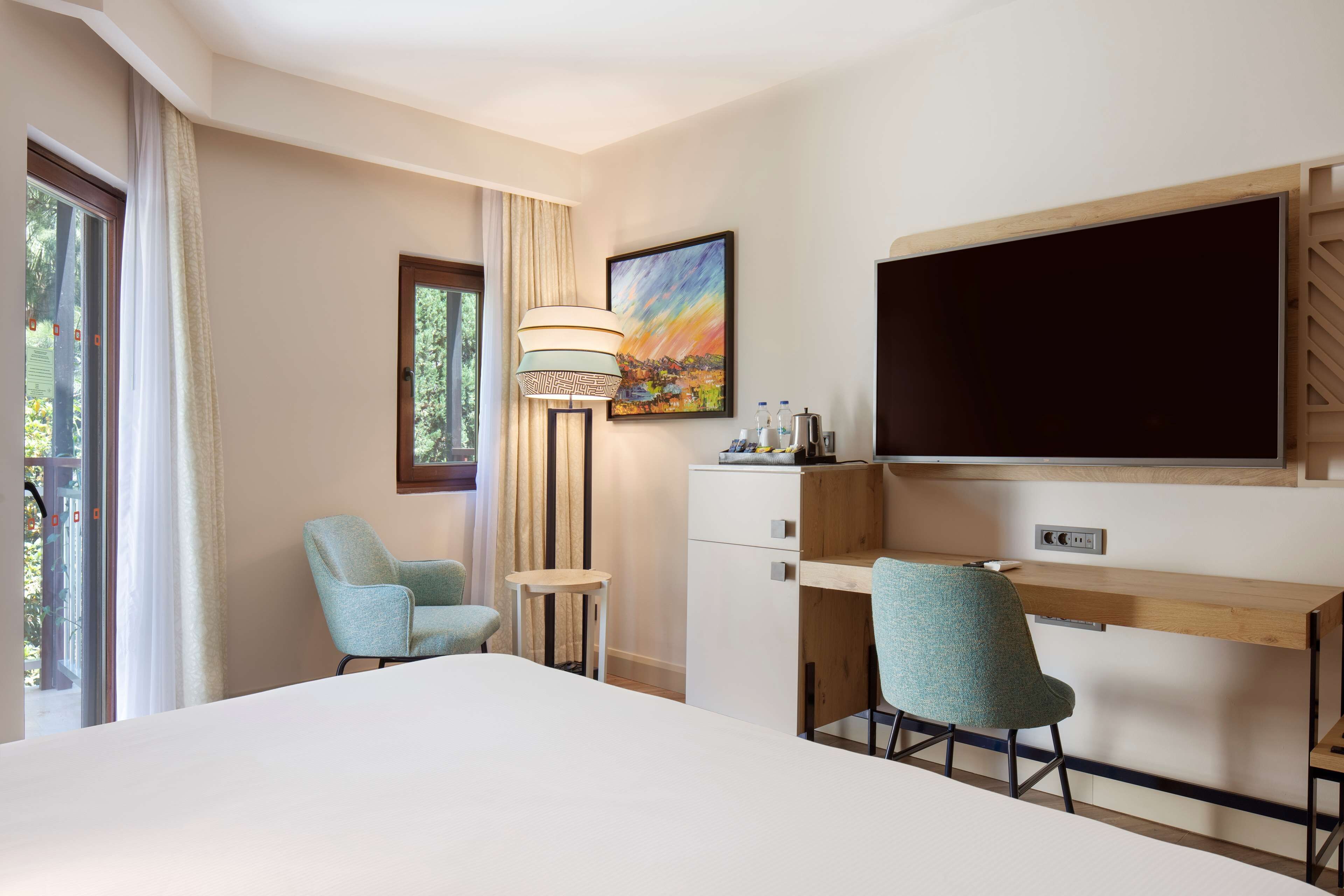 Habitación de hotel con cama, dos sillas, escritorio, lámpara, un cuadro y televisor.