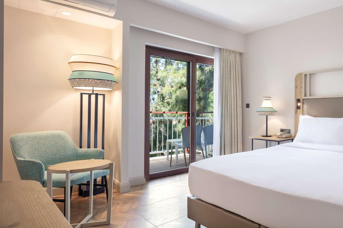 A hotel room with a bed, a chair, a table, a nightstand, and a balcony with a view of trees.