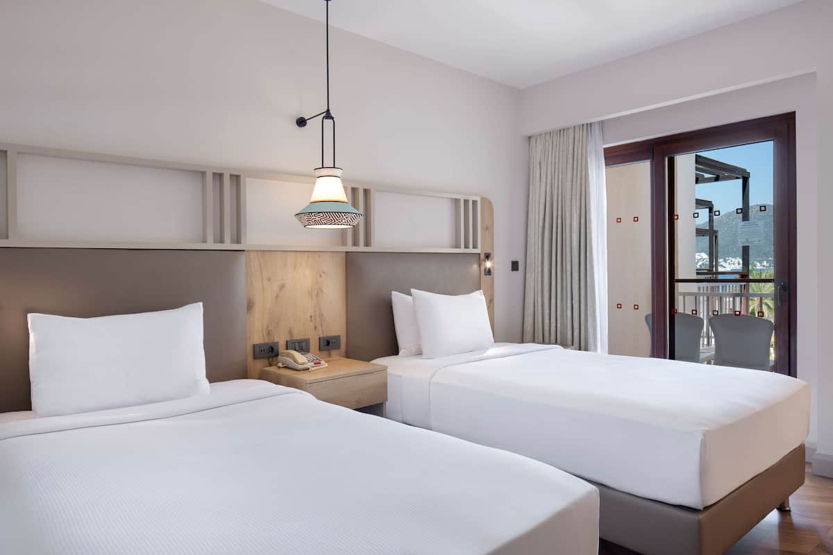 A hotel room with two beds, a wooden headboard, a bedside telephone, and a balcony with a view.