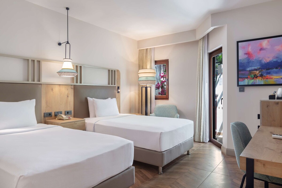 Habitación de hotel con dos camas, un escritorio, una silla, un televisor y un cuadro en la pared.