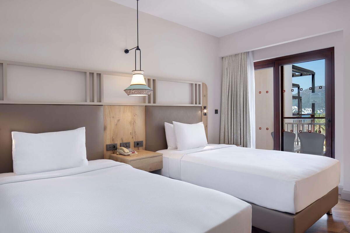 Habitación de hotel con dos camas, cabecera de madera, teléfono en la mesita de noche y balcón con vistas.