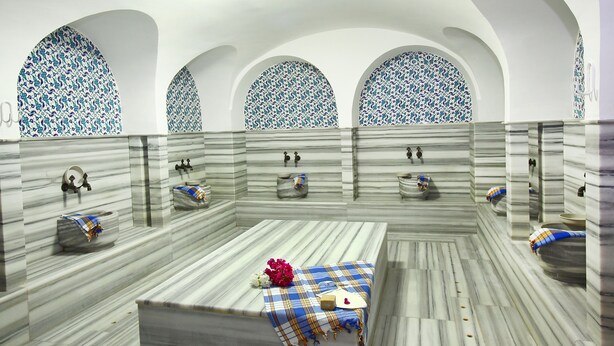 Casa de banho com três lavatórios, cada um com espelho e toalha. As paredes têm padrão marmorizado e há três janelas em arco com padrão azul e branco.