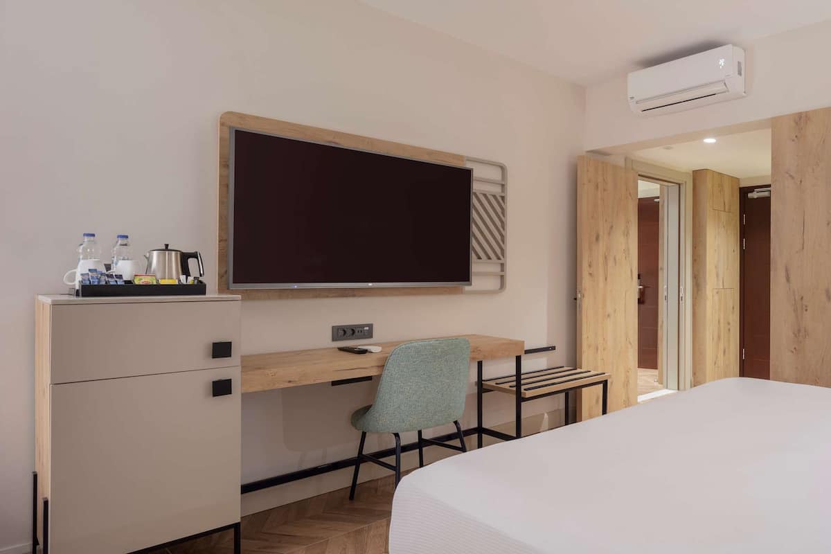 A hotel room with a bed, a desk with a chair, a flat-screen TV, and an air conditioning unit.