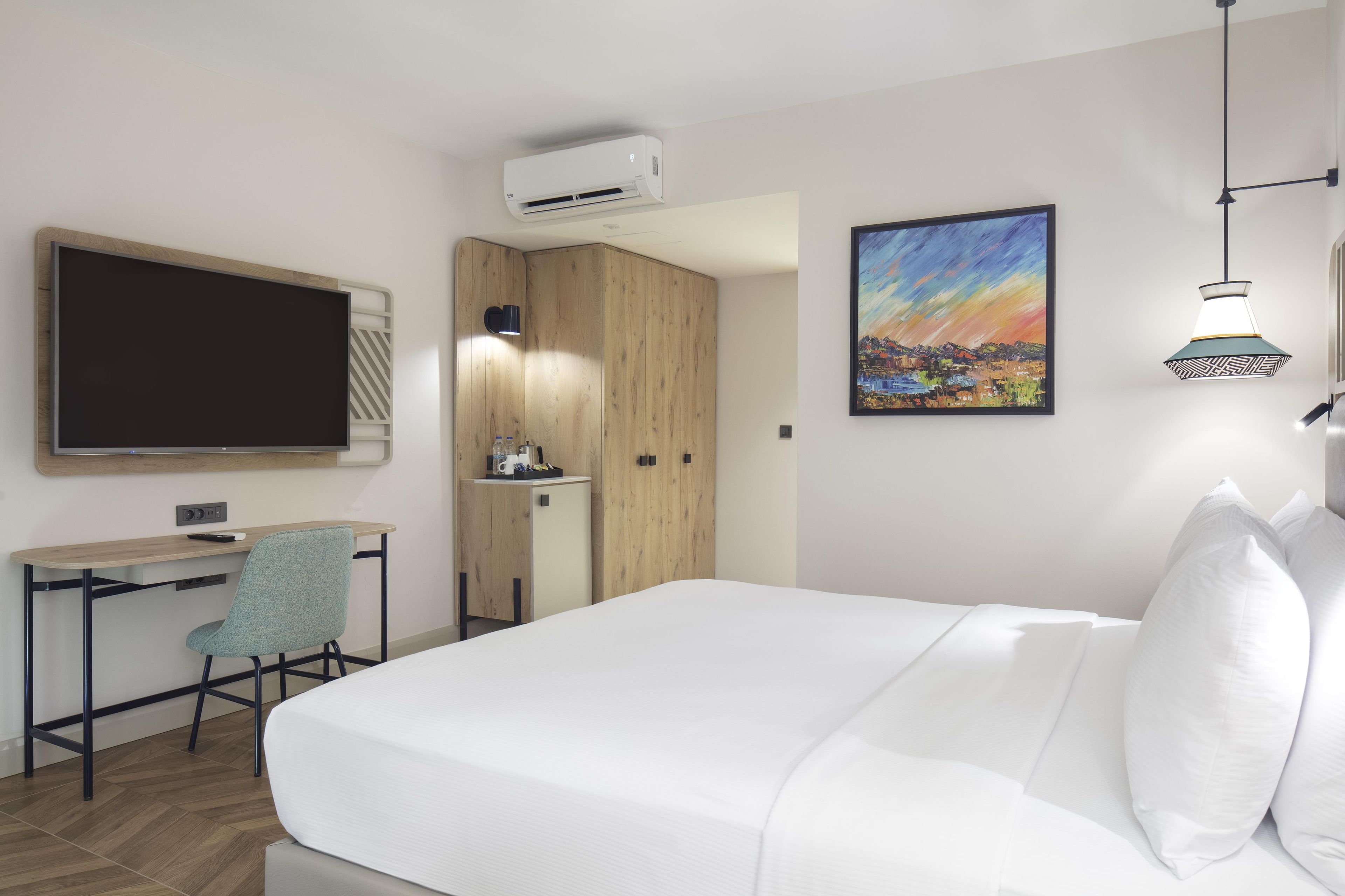 Habitación de hotel con cama, escritorio, silla, televisión, aire acondicionado, un cuadro y una lámpara.