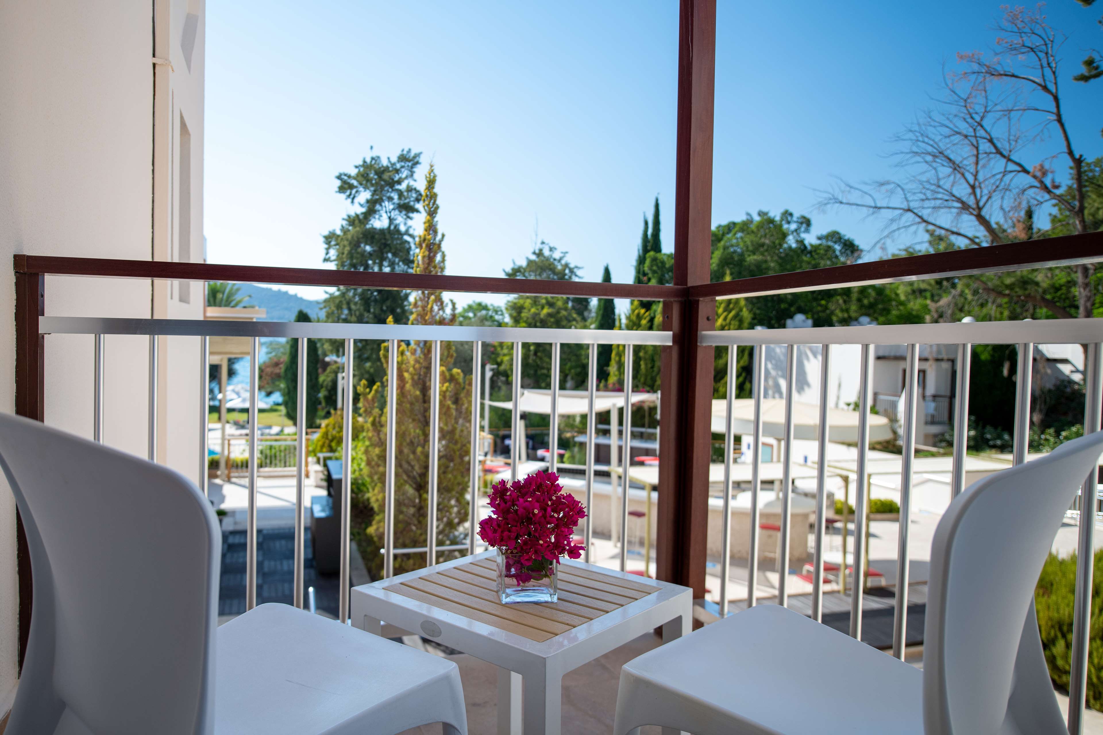 A balcony with two white chairs, a small table with a vase of flowers, and a view of a street with buildings and trees.