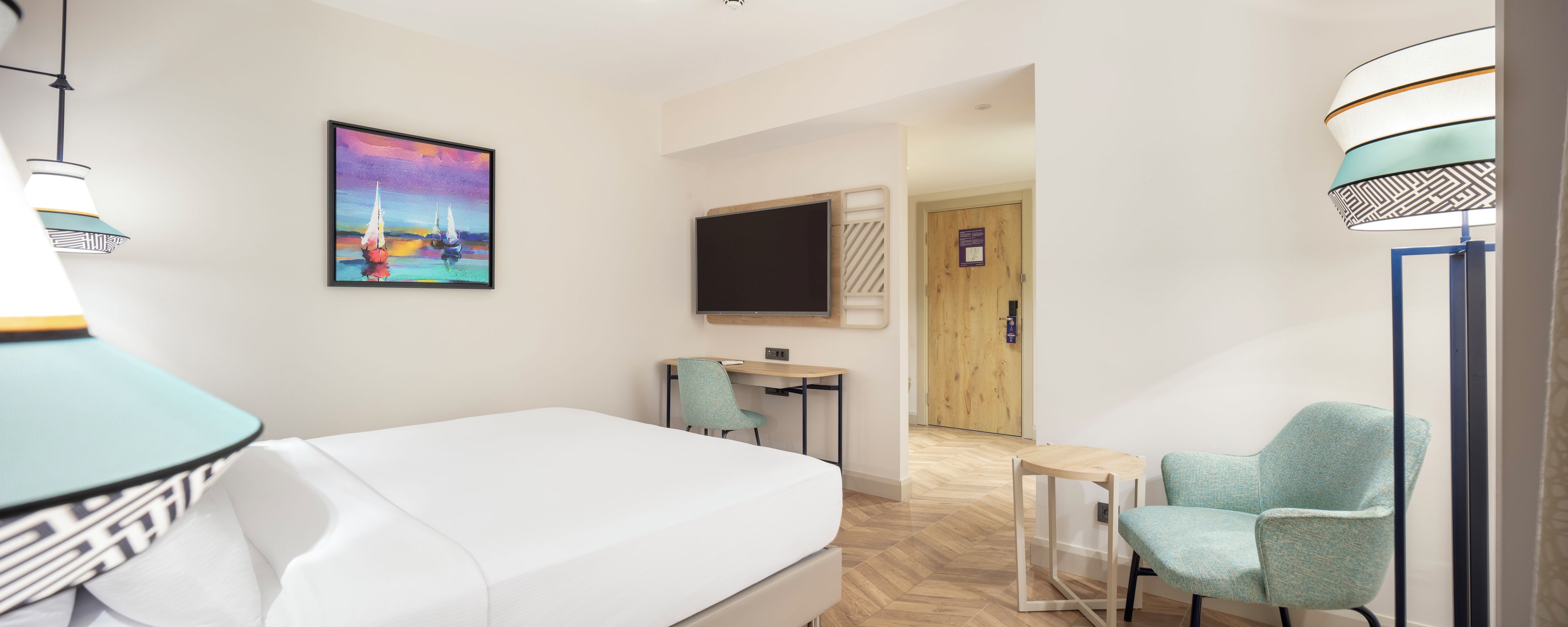 A hotel room with a bed, a desk, a chair, a TV, and a painting on the wall.