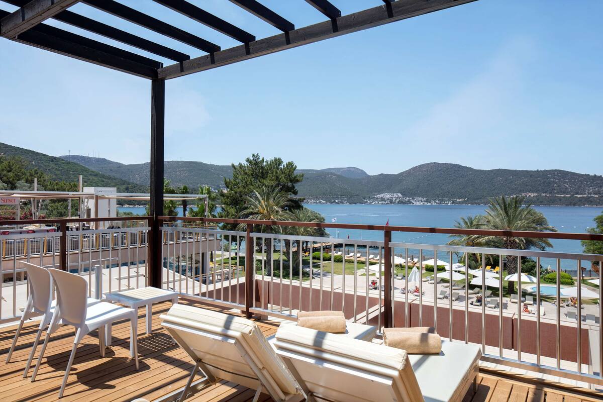 A balcony with white chairs and a table, overlooking a pool and mountains.