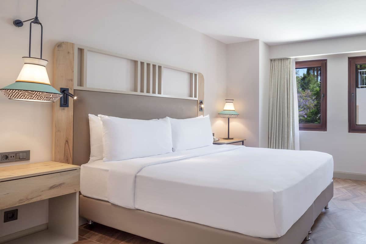 A hotel room with a bed, bedside table, lamp, chair, and a window with curtains.