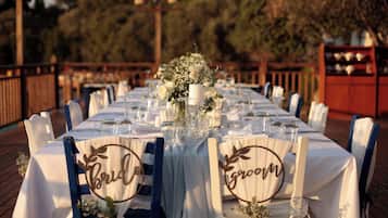 Una larga mesa de comedor dispuesta para una recepción de boda, con manteles blancos, cristalería y centros de mesa florales. Las sillas están etiquetadas con la palabra 'Bride' y 'Groom'.