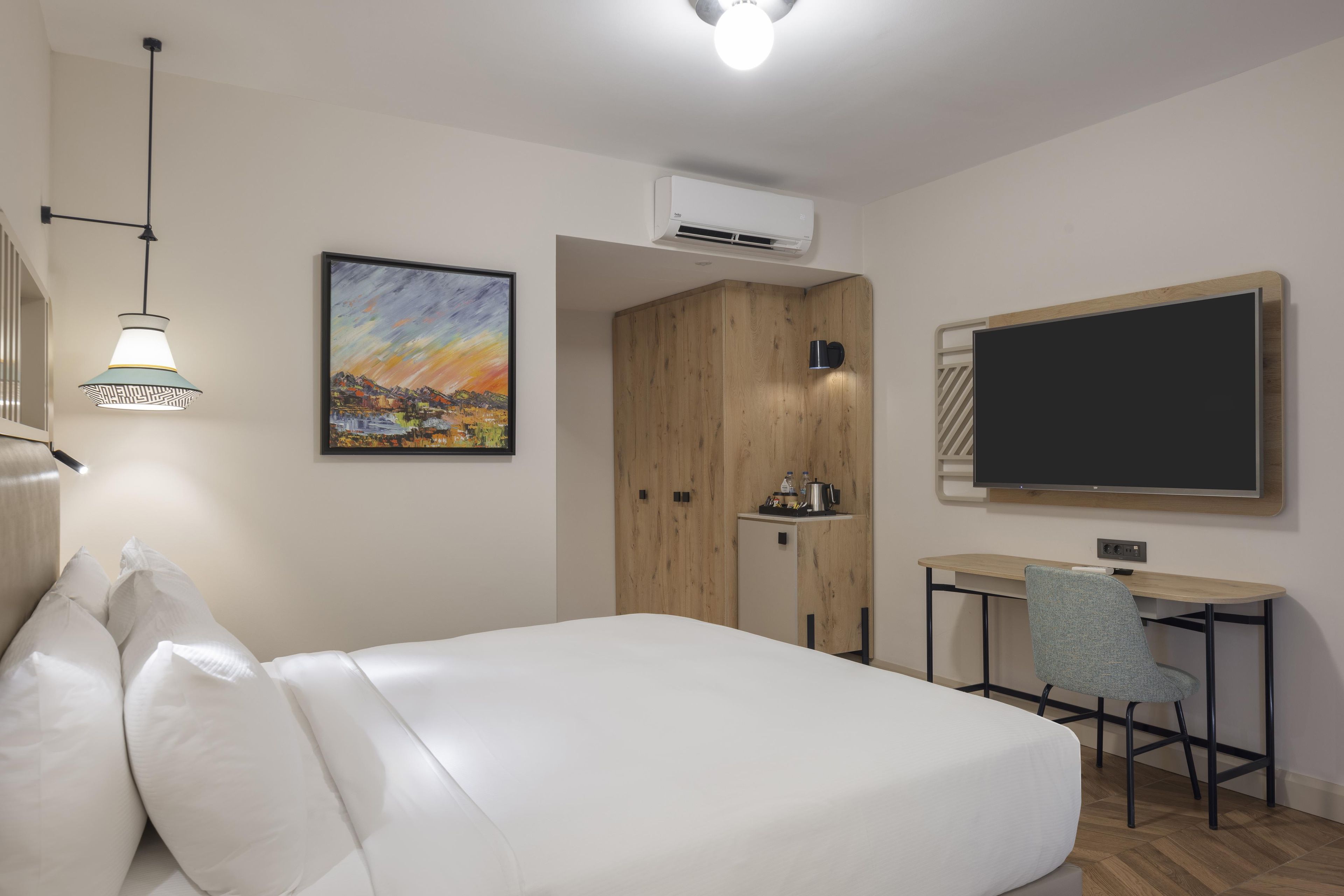 A hotel room with a bed, a desk, a chair, a painting, and an air conditioning unit.