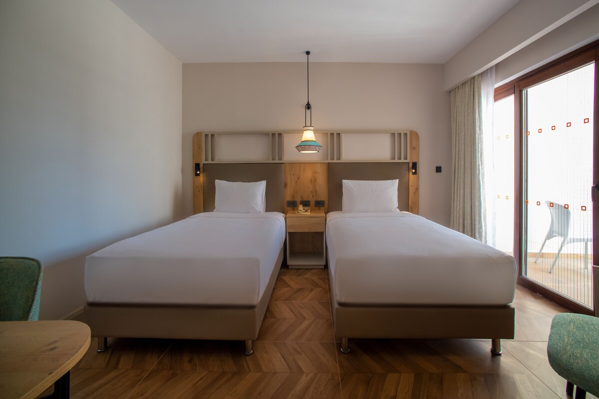 Quarto de hotel com duas camas de solteiro, cabeceira de madeira, uma mesinha e vista para o exterior através de uma janela grande.