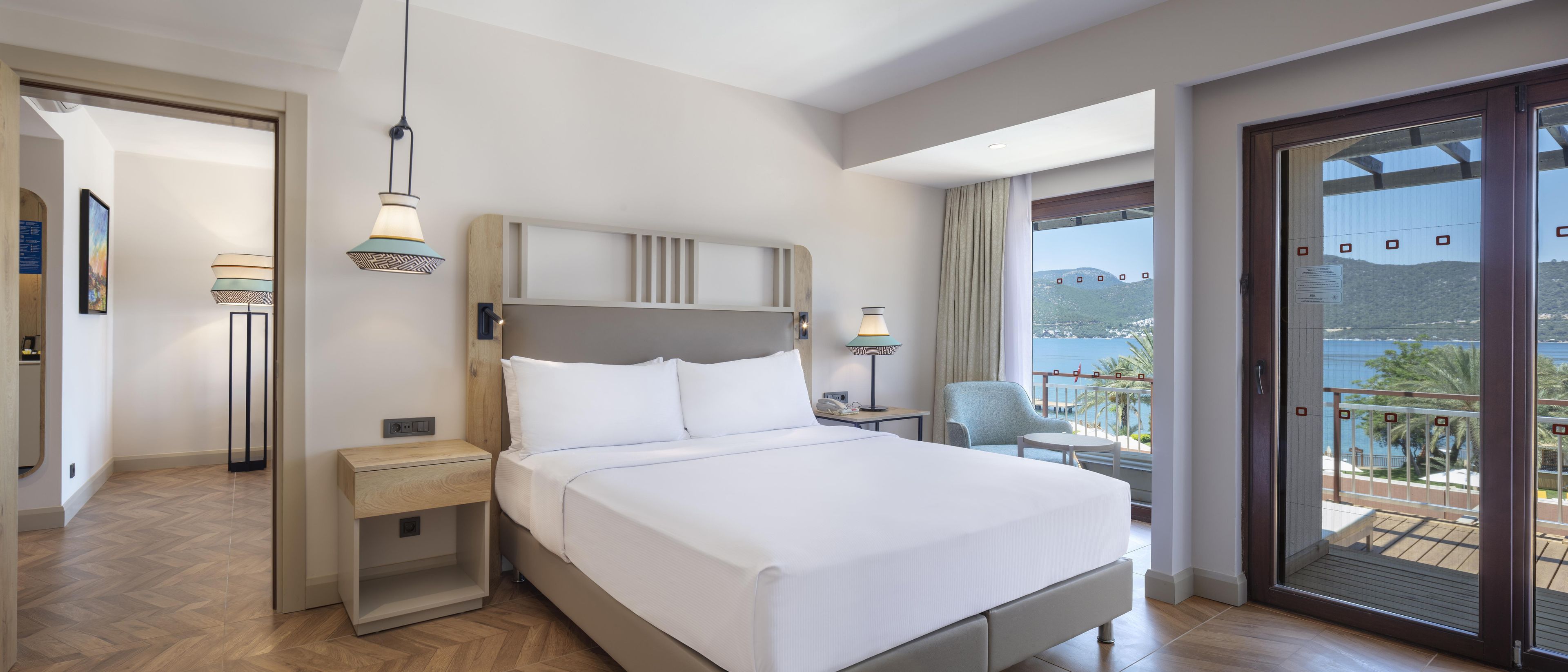 A hotel room with a bed, bedside tables, a chair, a balcony with a view of the sea, and a door.