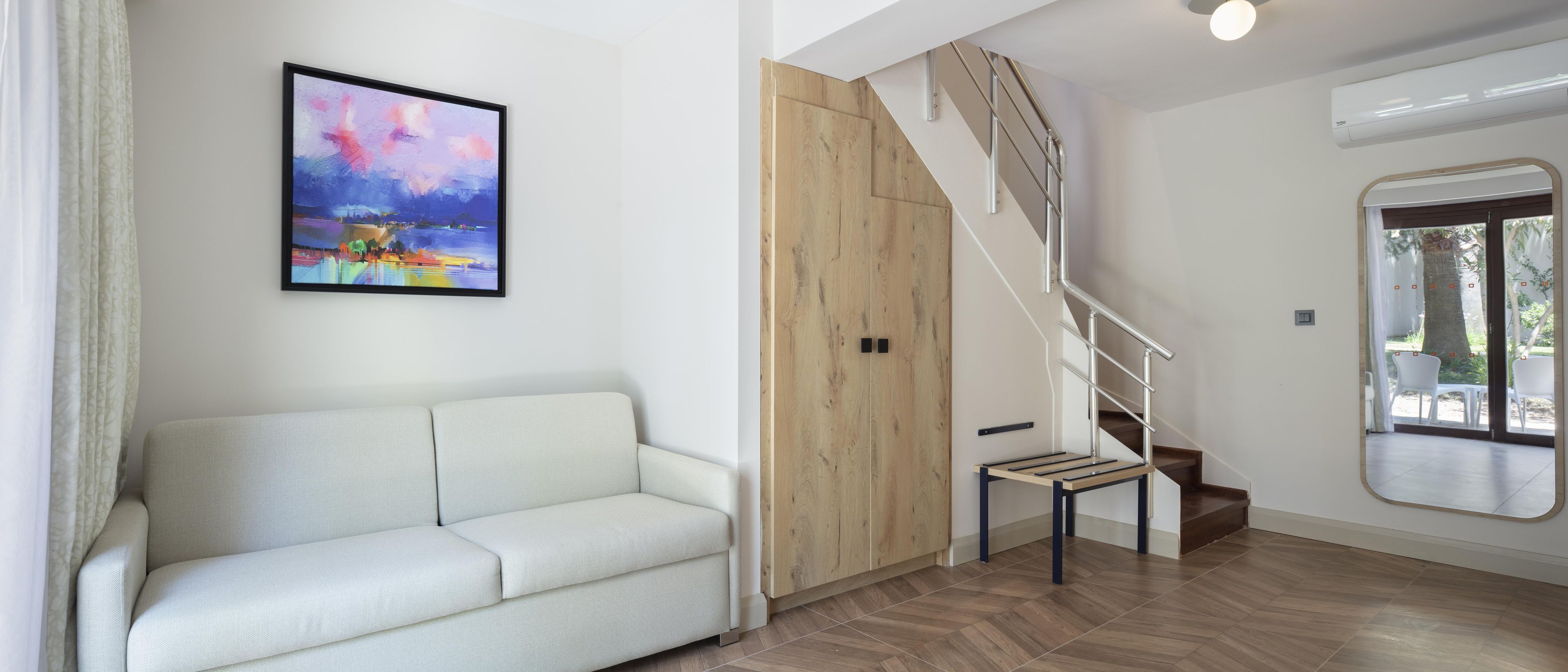 Un interior modern amb un sofà blanc, una porta de fusta, una escala amb barana metàl·lica i una finestra amb vista a vegetació.
