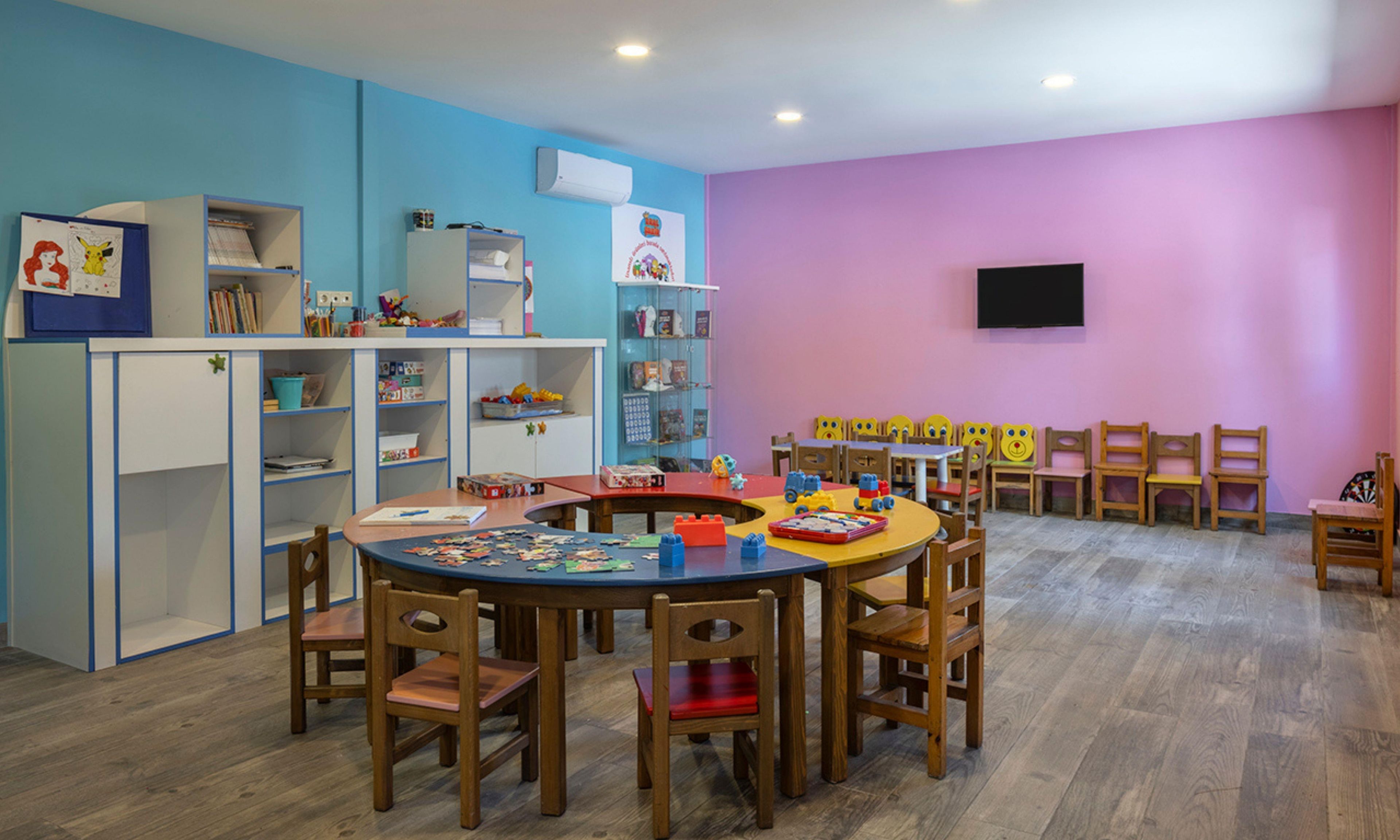 Ein Spielzimmer für Kinder mit einem runden Tisch, Stühlen, Regalen voller Spielzeug und einem Fernseher an der Wand.