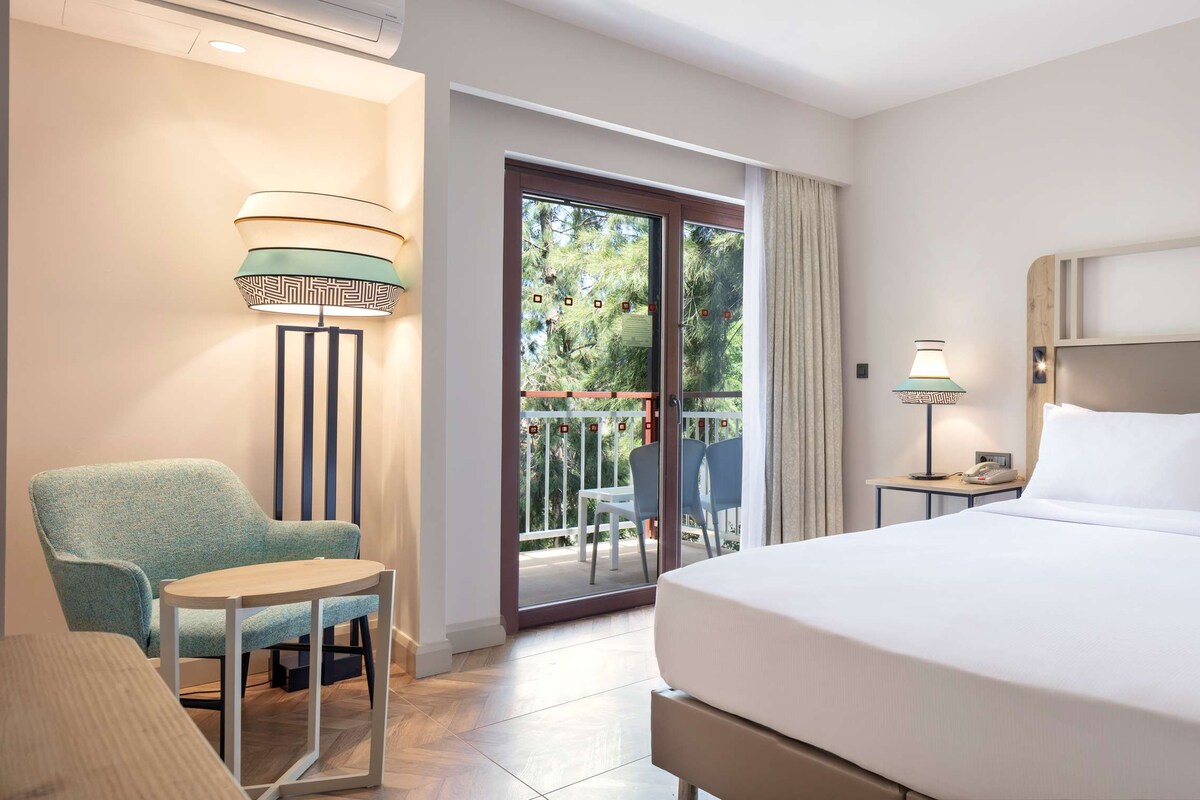 Habitación de hotel con cama, una silla, una mesa, mesita de noche y balcón con vistas a árboles.
