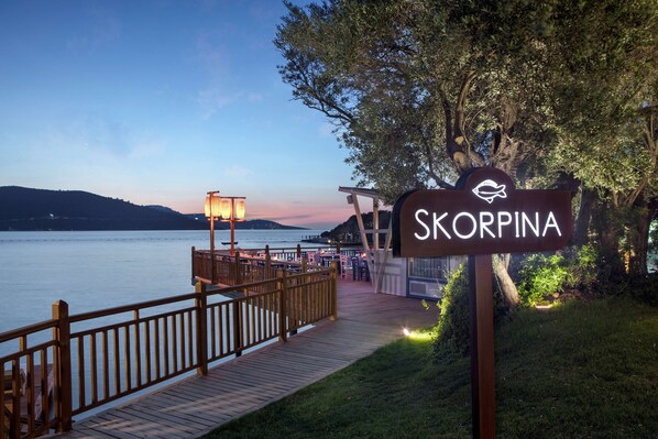 A wooden deck leading to a waterfront restaurant named Skorpina, with a view of the water and distant hills. - DoubleTree by Hilton Bodrum Işıl Club Ultra All Inclusive Resort (Bodrum)