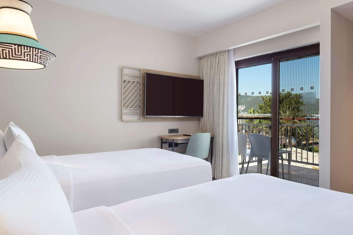 A hotel room with two beds, a TV, a small table with chairs, a balcony with a view, and a minibar.