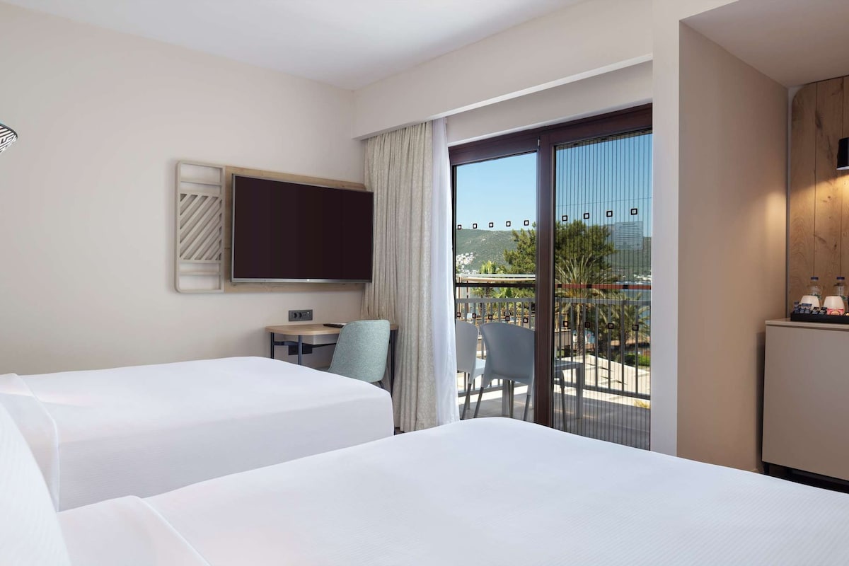 A hotel room with two beds, a TV, a small table with chairs, a balcony with a view, and a minibar.