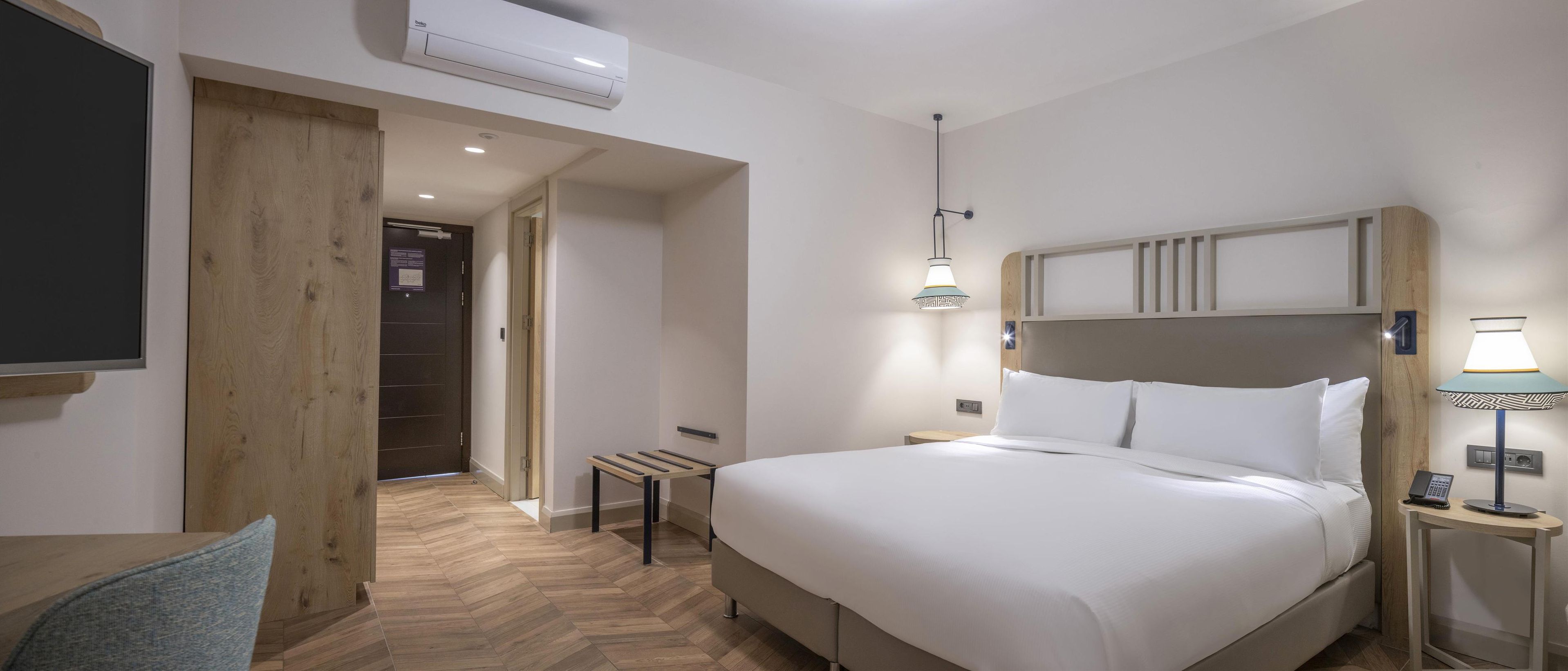 Una habitación de hotel con una cama grande, una mesita de noche con lámpara, un televisor montado en la pared y una puerta de madera.