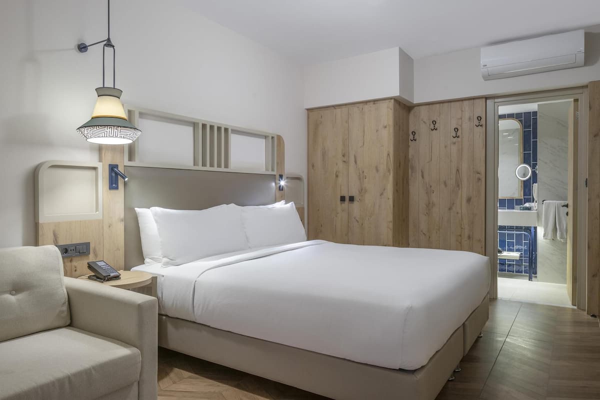 Ett hotellrum med en säng, ett sängbord med en lampa, en soffa, en garderob i trä och ett badrum som syns genom en öppen dörr.