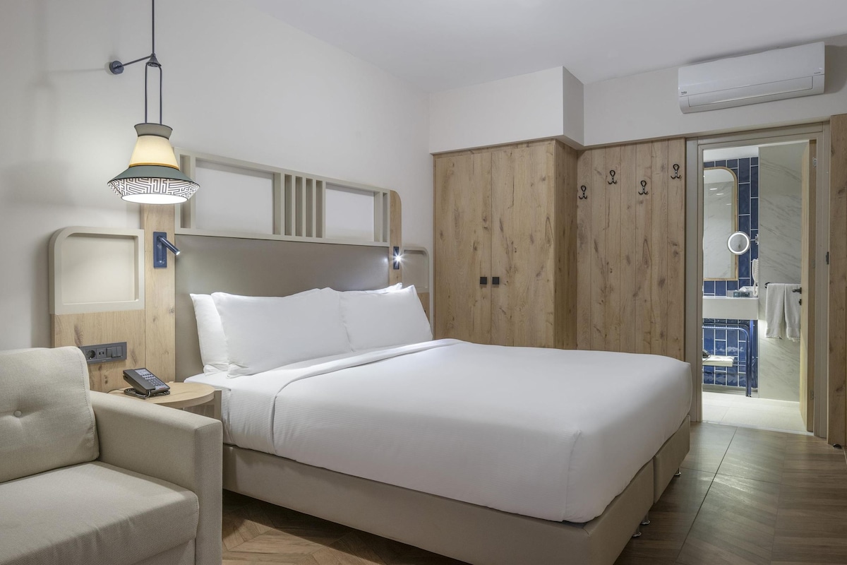 Habitación de hotel con una cama, una mesita de noche con lámpara, un sofá, un armario de madera y un baño visible a través de una puerta abierta.