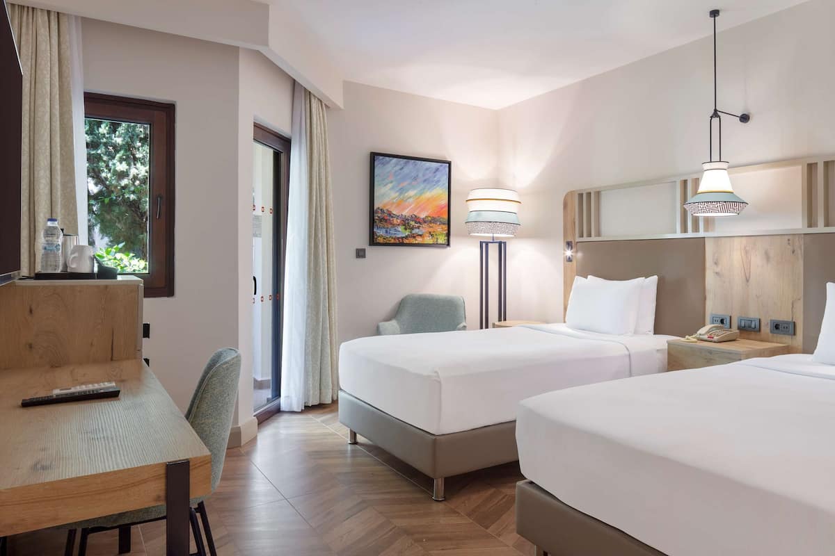 A hotel room with two beds, a desk, a chair, a TV, a painting, and a lamp.