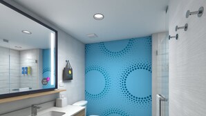 Un baño moderno con un lavabo blanco, tocador de madera y un inodoro. Hay un espejo sobre el lavabo y un cubo de basura rojo al lado del inodoro. Las paredes están decoradas con un estampado azul a lunares.