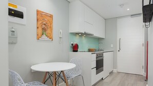 Een kleine keuken met witte kastjes, een fornuis en een spoelbak. Er staat een witte tafel met twee stoelen en een schilderij aan de muur.