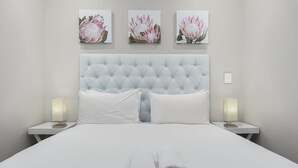 Een netjes opgemaakt bed met witte lakens en twee opgerolde handdoeken aan het voeteneinde. Aan weerszijden van het bed staan twee nachtkastjes met lampen. Boven het bed hangen drie ingelijste bloemwerken op de muur.