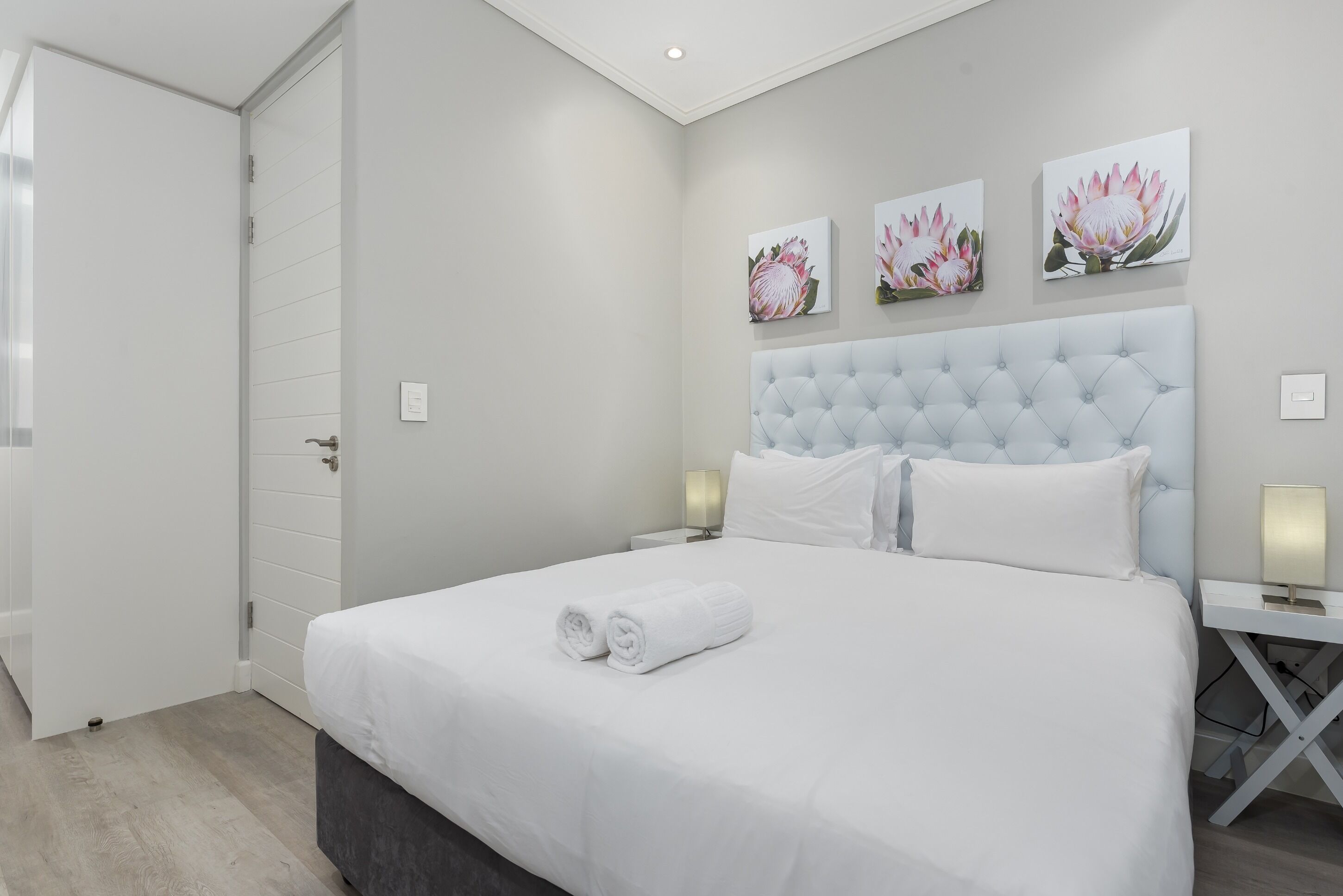 A neatly made bed with white linens and rolled towels, a tufted headboard, and three wall-mounted floral paintings.