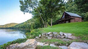 A rustic cabin with a stone foundation, surrounded by a lush green landscape and a body of water. - SECLUDED WHITE RIVERFRONT CABIN -SLEEPS 2-10, GREAT FAMILY & FISHERMEN GETAWAY (Norfork)