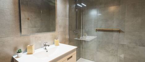 Un baño moderno con un espejo grande, un lavamanos blanco, una ducha y un estante pequeño.