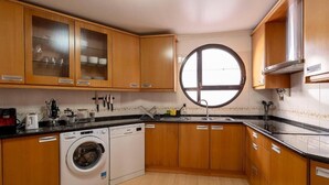 A compact kitchen with wooden cabinets, a built-in washing machine, and a large window. - Ericeira Paradise House & Suites (Mafra)