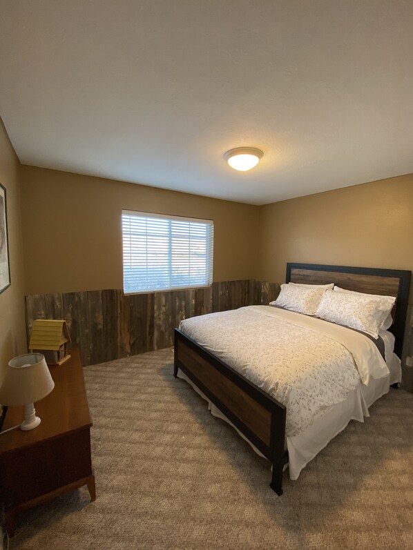 A bedroom with a bed, a nightstand, a lamp, a window with blinds, and a ceiling light. - Two Bit Ranch House (Escalante)