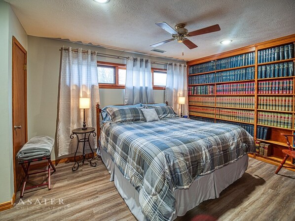 Une chambre à coucher avec un grand lit, une bibliothèque en bois, un ventilateur de plafond et une fenêtre avec des rideaux.