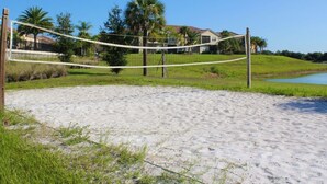 A sandy beach with a volleyball net, palm trees, and a house in the background.