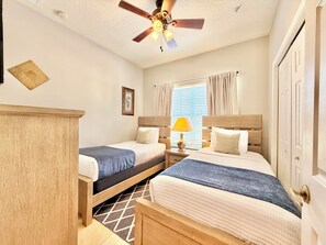 A hotel room with two beds, a ceiling fan, a window with curtains, and a wooden nightstand with a lamp.