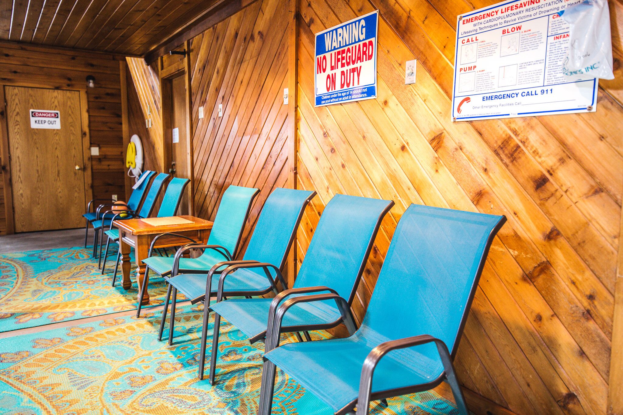 A room with wooden walls and a patterned carpet, featuring blue chairs and a table. A warning sign indicates no lifeguard on duty.