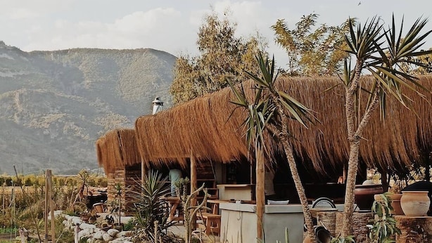 A rustic thatched-roof structure with a stone bench and a small table, surrounded by rocks and sparse vegetation, with mountains in the background. - Bhavana Gokova (Ula)
