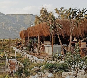 A rustic thatched-roof structure with a stone bench and a small table, surrounded by rocks and sparse vegetation, with mountains in the background. - Bhavana Gokova (Ula)