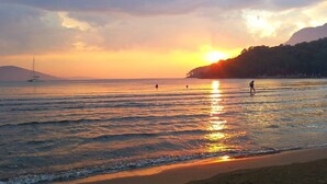A sunset over a beach with calm waters, a sailboat, and a person walking on the shore. - Bhavana Gokova (Ula)